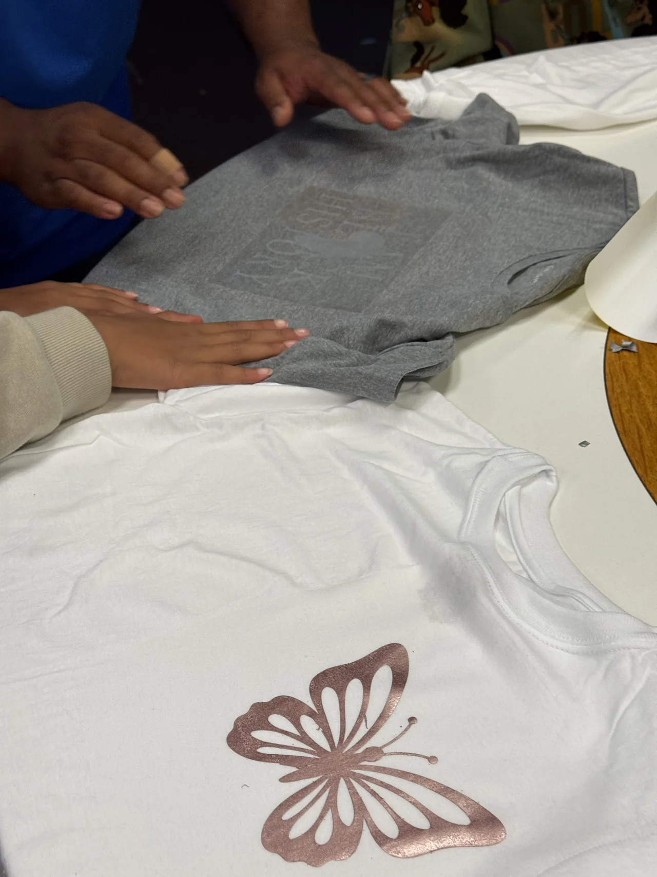 Students smooth a gray shirt beside a finished white shirt printed with a butterfly.