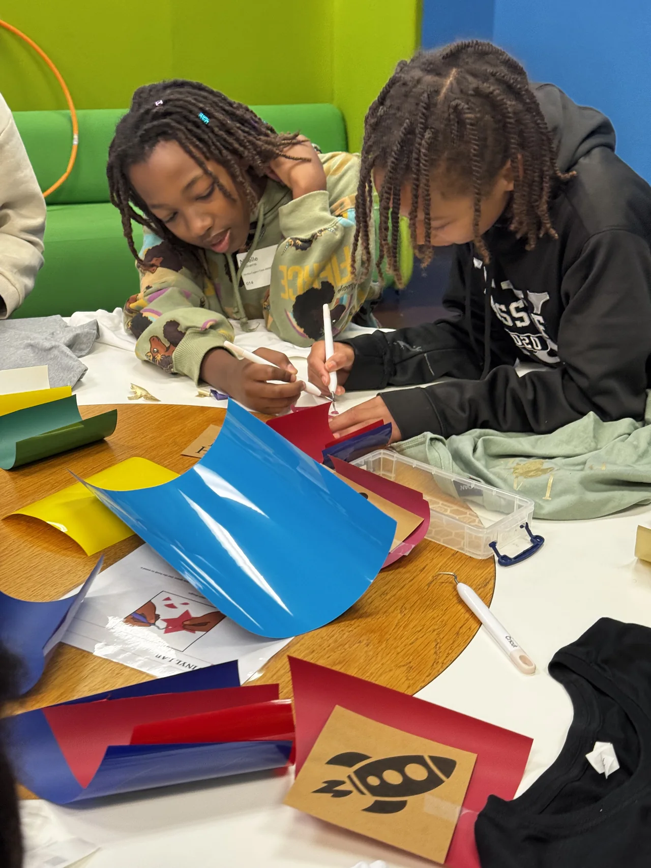 Two students work side by side on their shirt designs at a table covered with vinyl and stencils.
