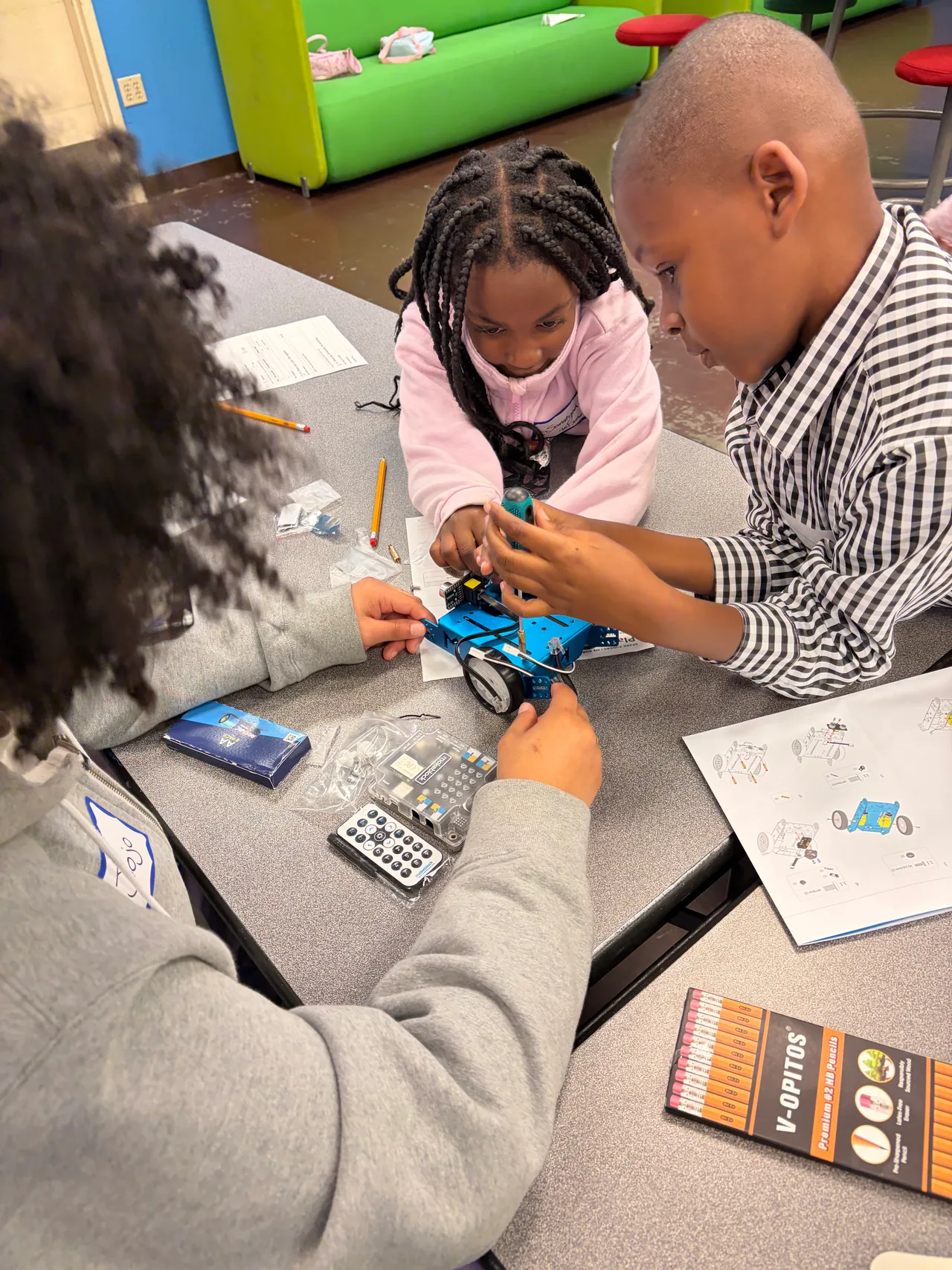 Two students and an adult work together on a blue mBot, one holding the robot steady while another turns a screwdriver.