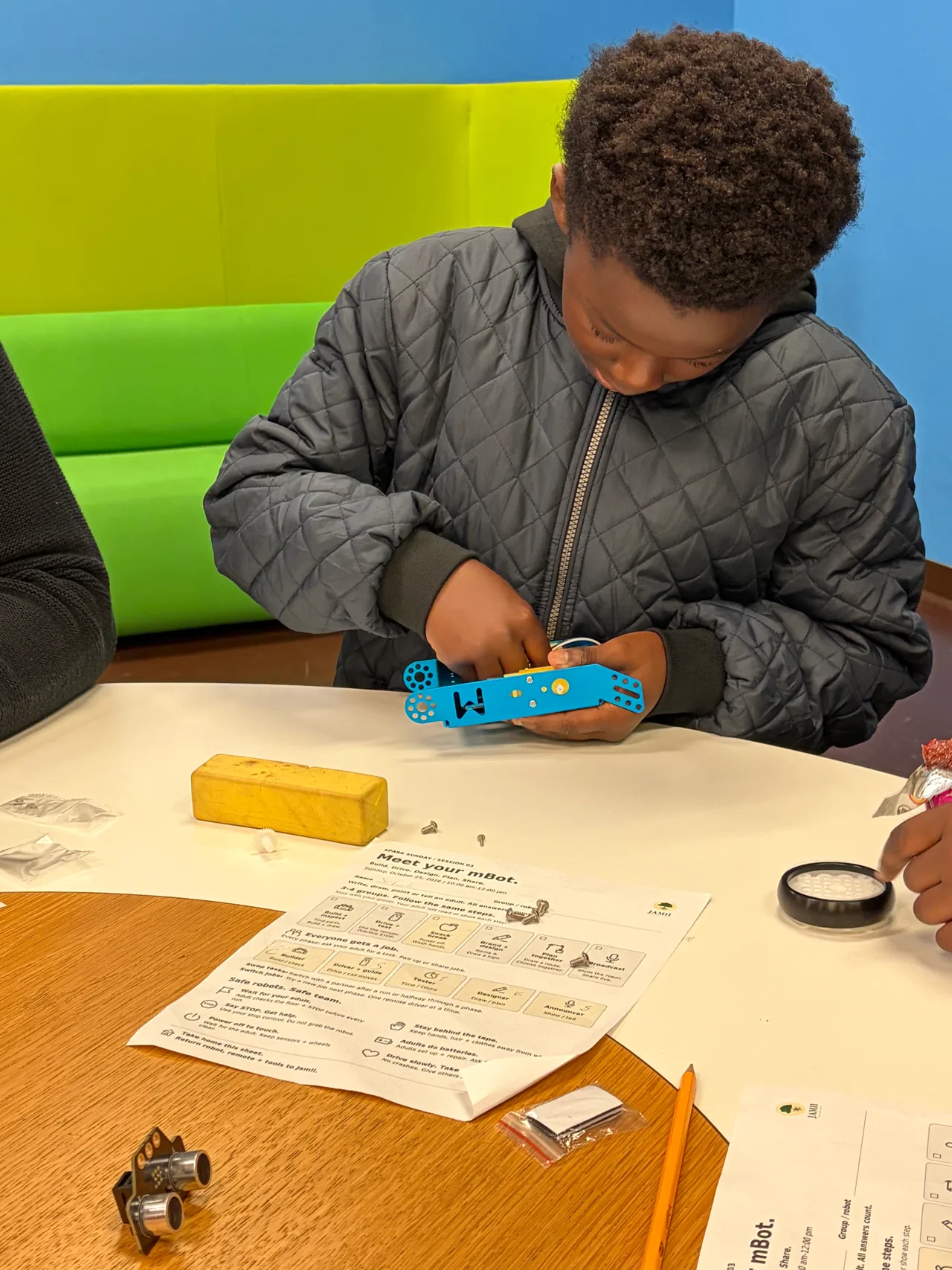 A student fits parts into a blue robot chassis beside a loose wheel, an ultrasonic sensor and illustrated instructions.
