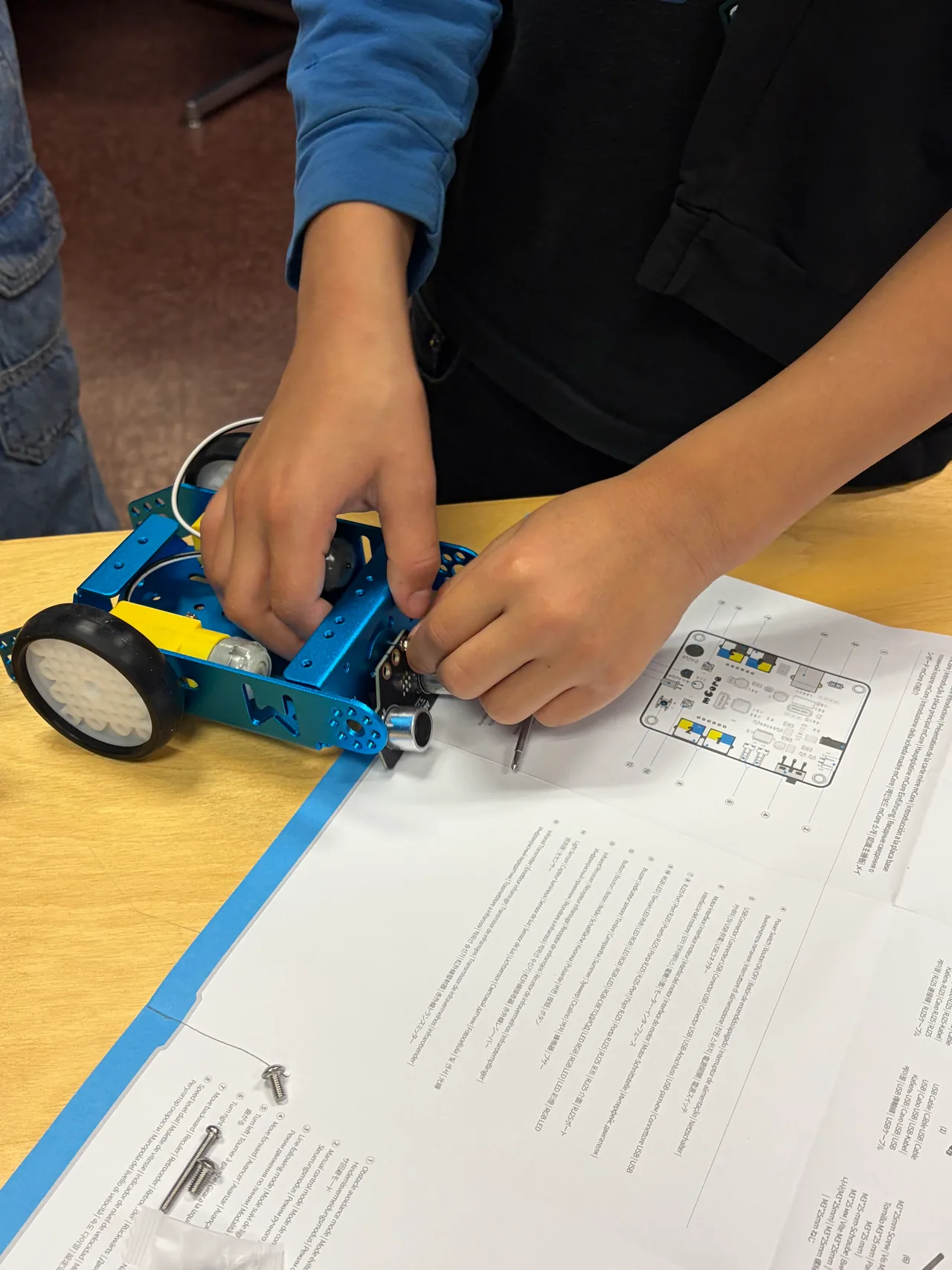 Close view of a student connecting components inside a wheeled blue mBot over an open assembly booklet.