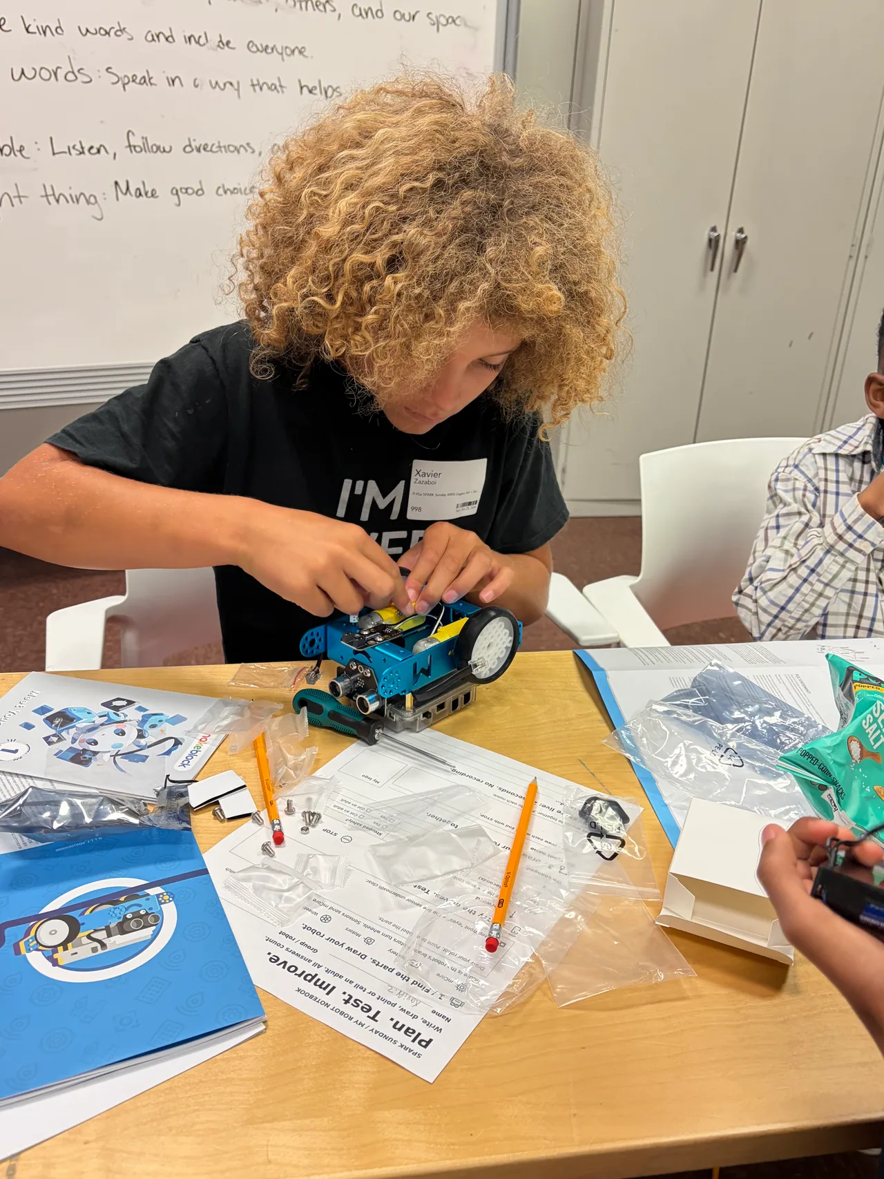 A student concentrates on the top of a blue mBot surrounded by instruction sheets, tools and kit packaging.