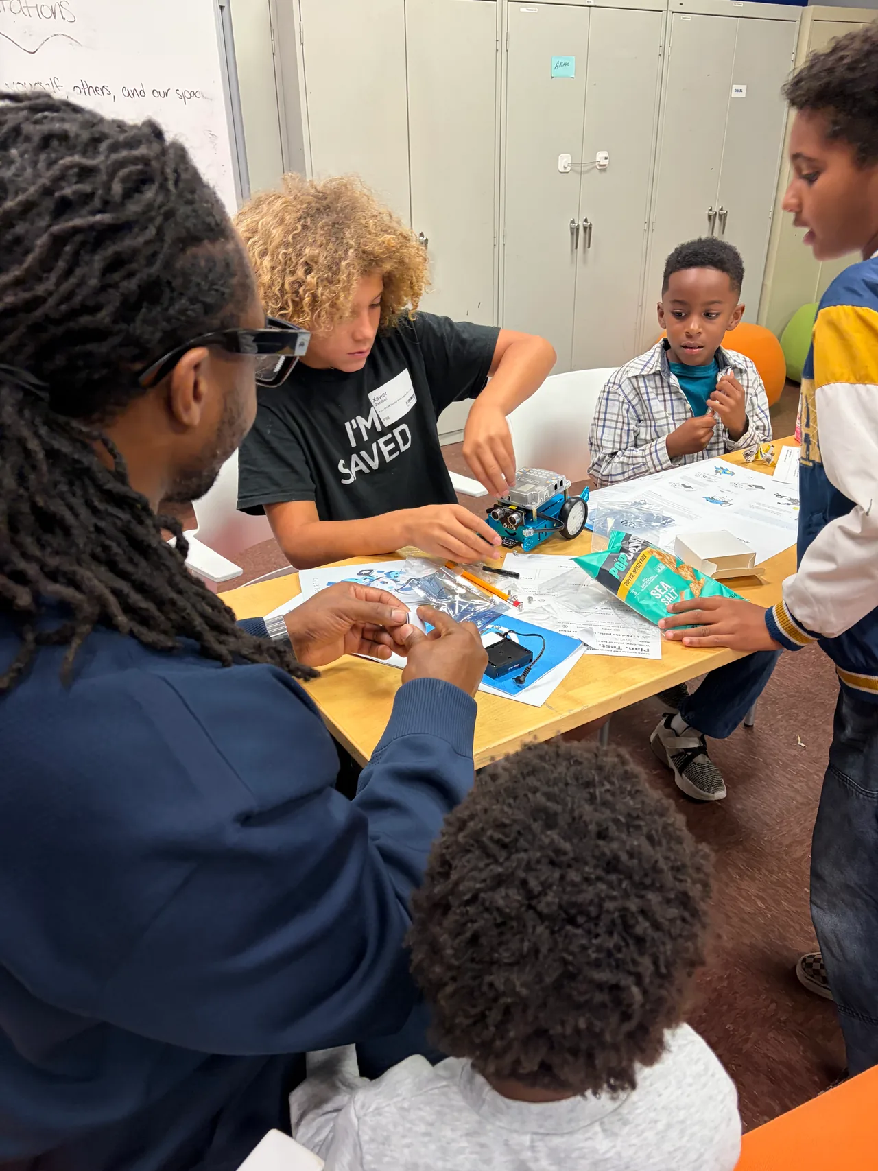Students gather around a blue robot at a shared table while an adult helps prepare a component.