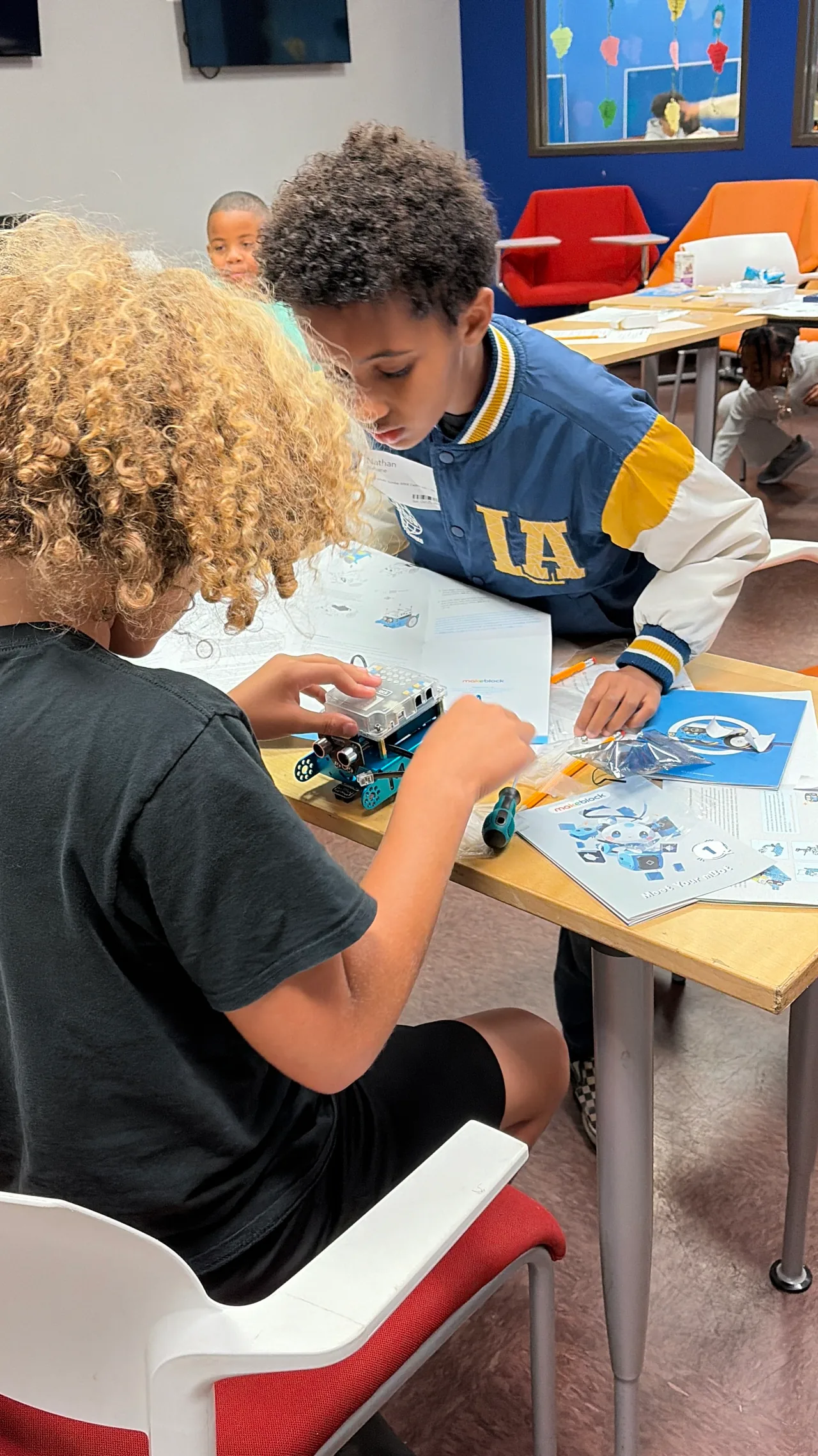 Two students lean over a blue mBot and an open illustrated Makeblock booklet as they work through the assembly.