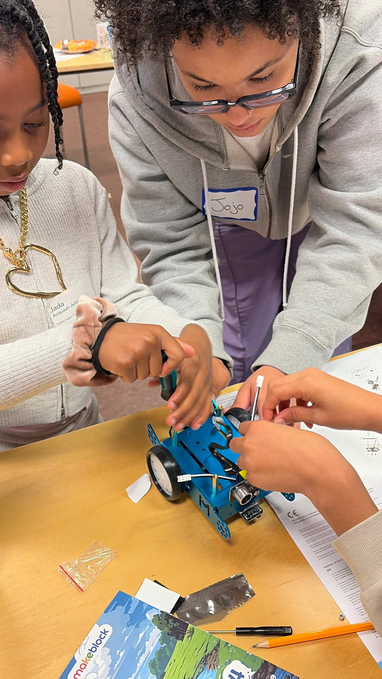 A student uses a screwdriver on a blue robot while an adult and another participant help steady the build.
